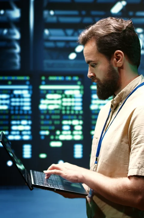 Man working on a laptop in front of a large digital display with server data.