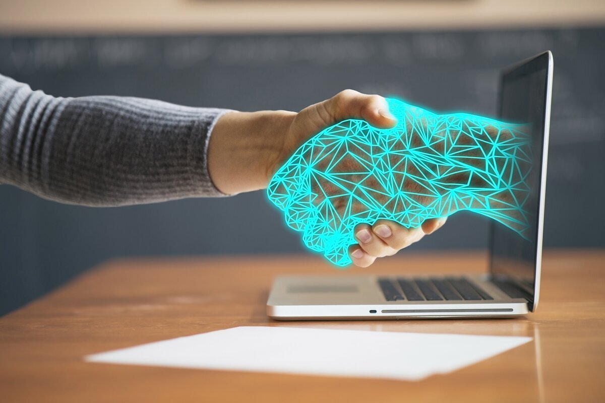 A person’s hand shaking a glowing digital hand emerging from a laptop screen